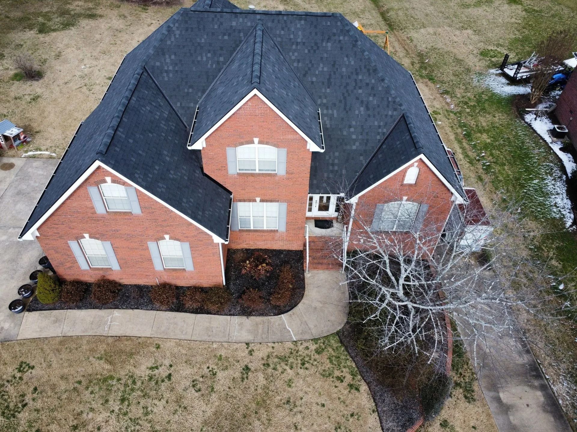 An aerial view of a two-story brick house with a dark shingled roof, light shutters, and a concrete walkway.