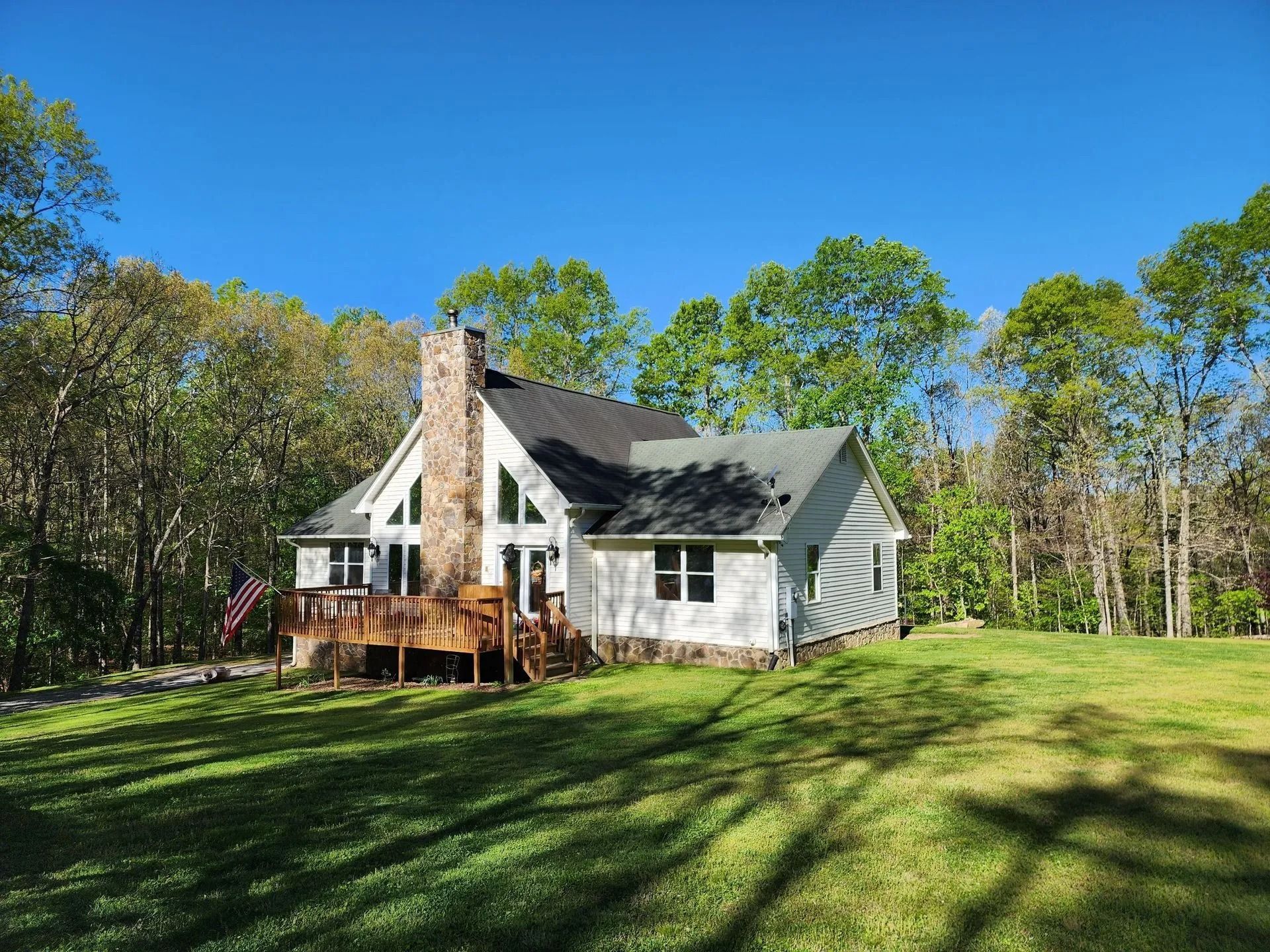 A white house with a stone chimney and wooden deck sits in a sunny, green yard surrounded by trees under a clear blue sky.