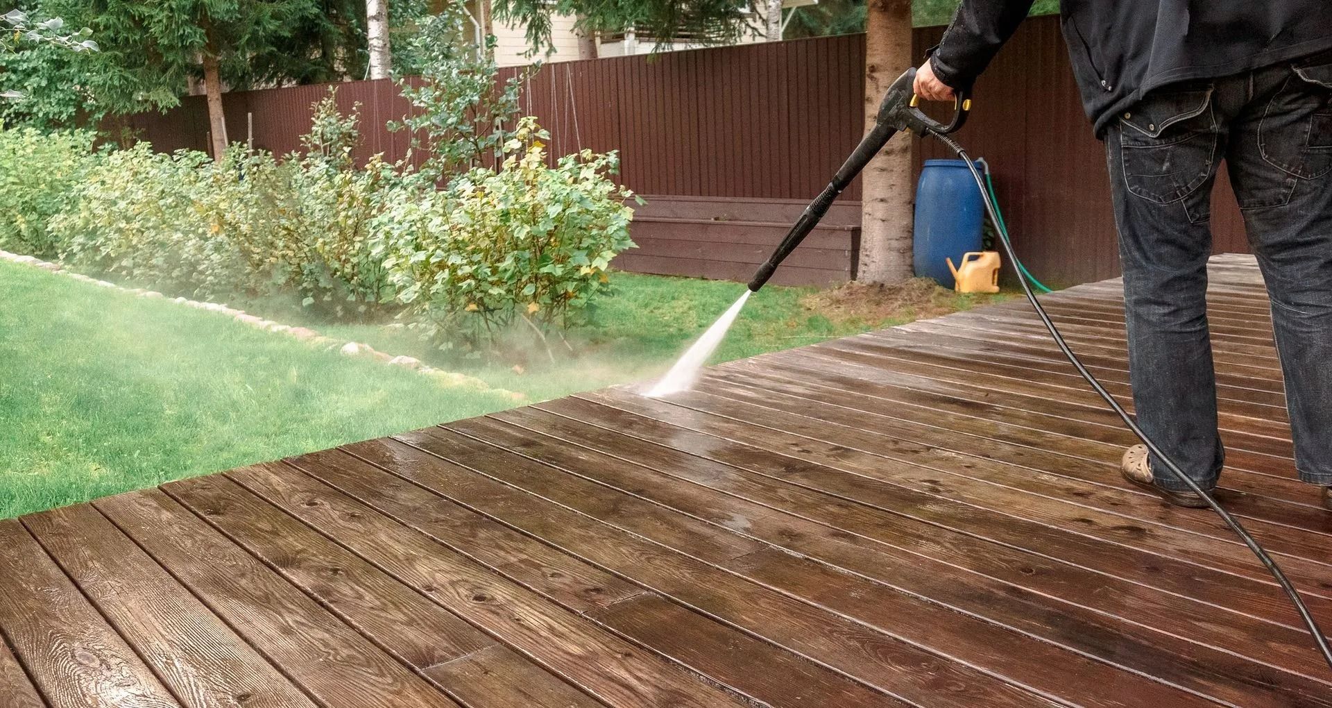 Person pressure-washing a wooden deck outdoors with a spray wand, water spraying across the boards