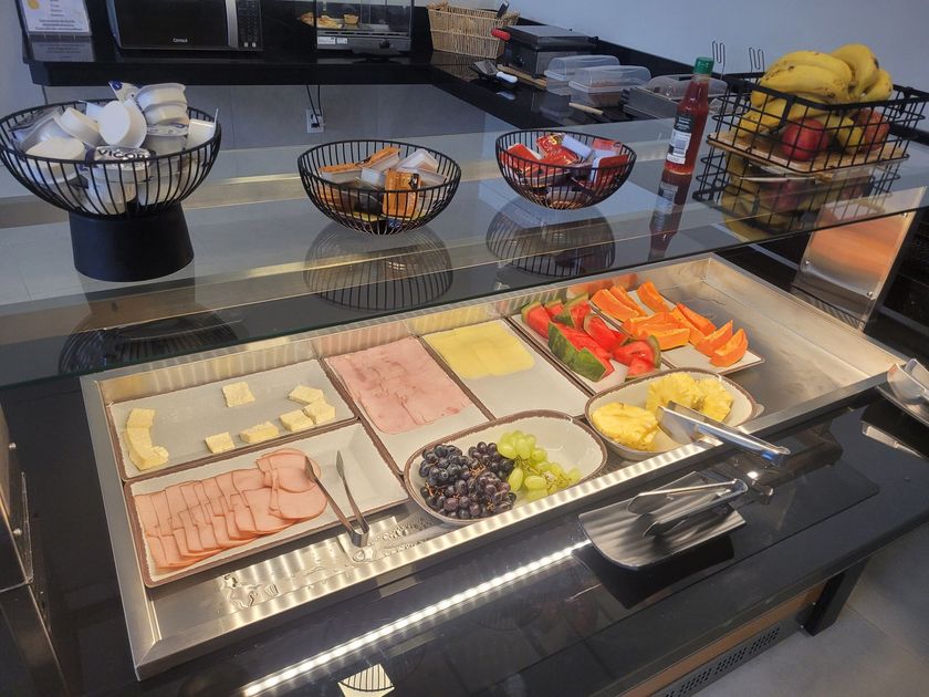 Buffet de café da manhã com tigelas de frutas, doces e frios fatiados em um balcão de vidro.