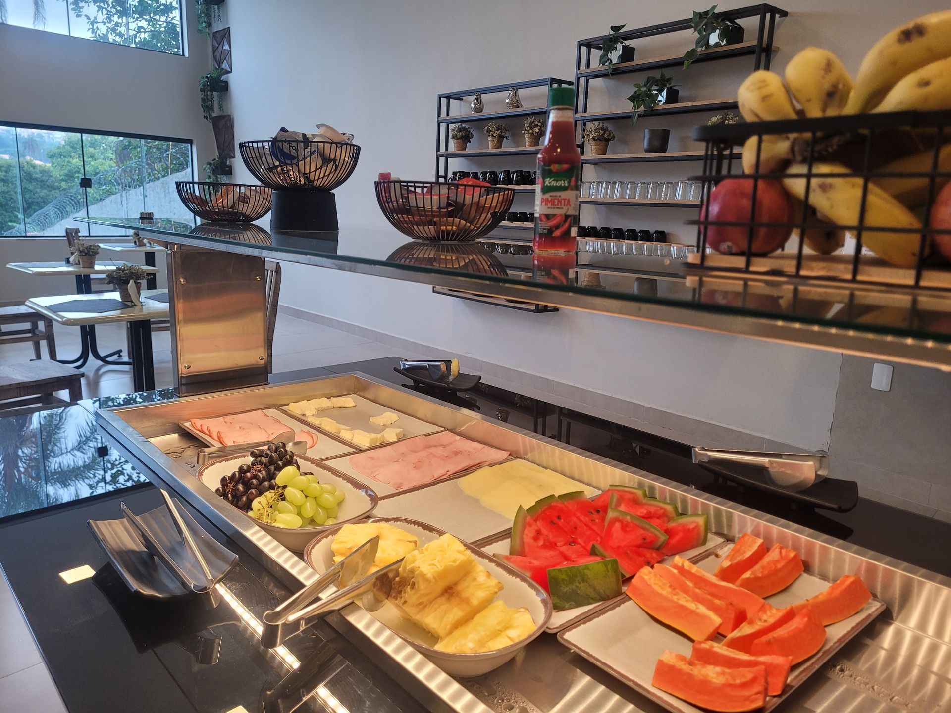 Balcão de buffet com bandejas de frutas, salmão fatiado e itens variados de café da manhã em uma área de refeições bem iluminada.