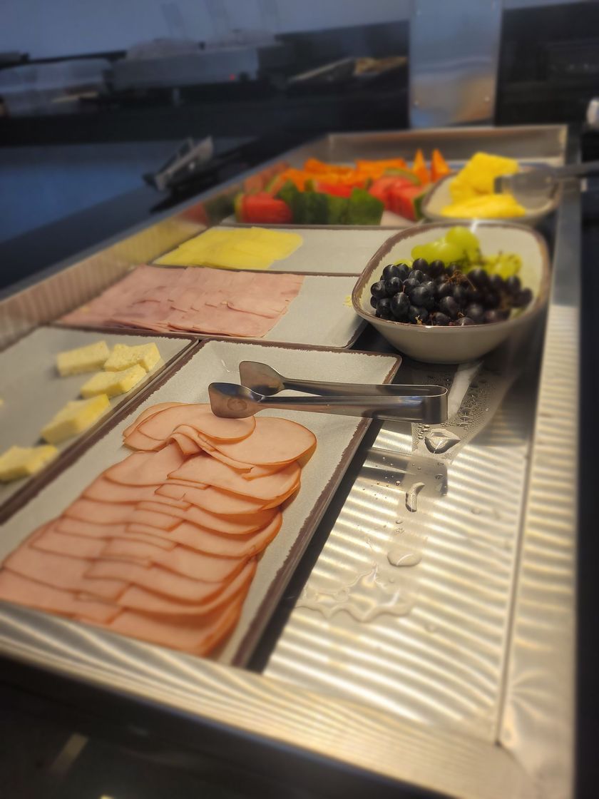 Buffet de saladas com frios fatiados, azeitonas, queijos e vegetais coloridos em bandejas de aço inoxidável.