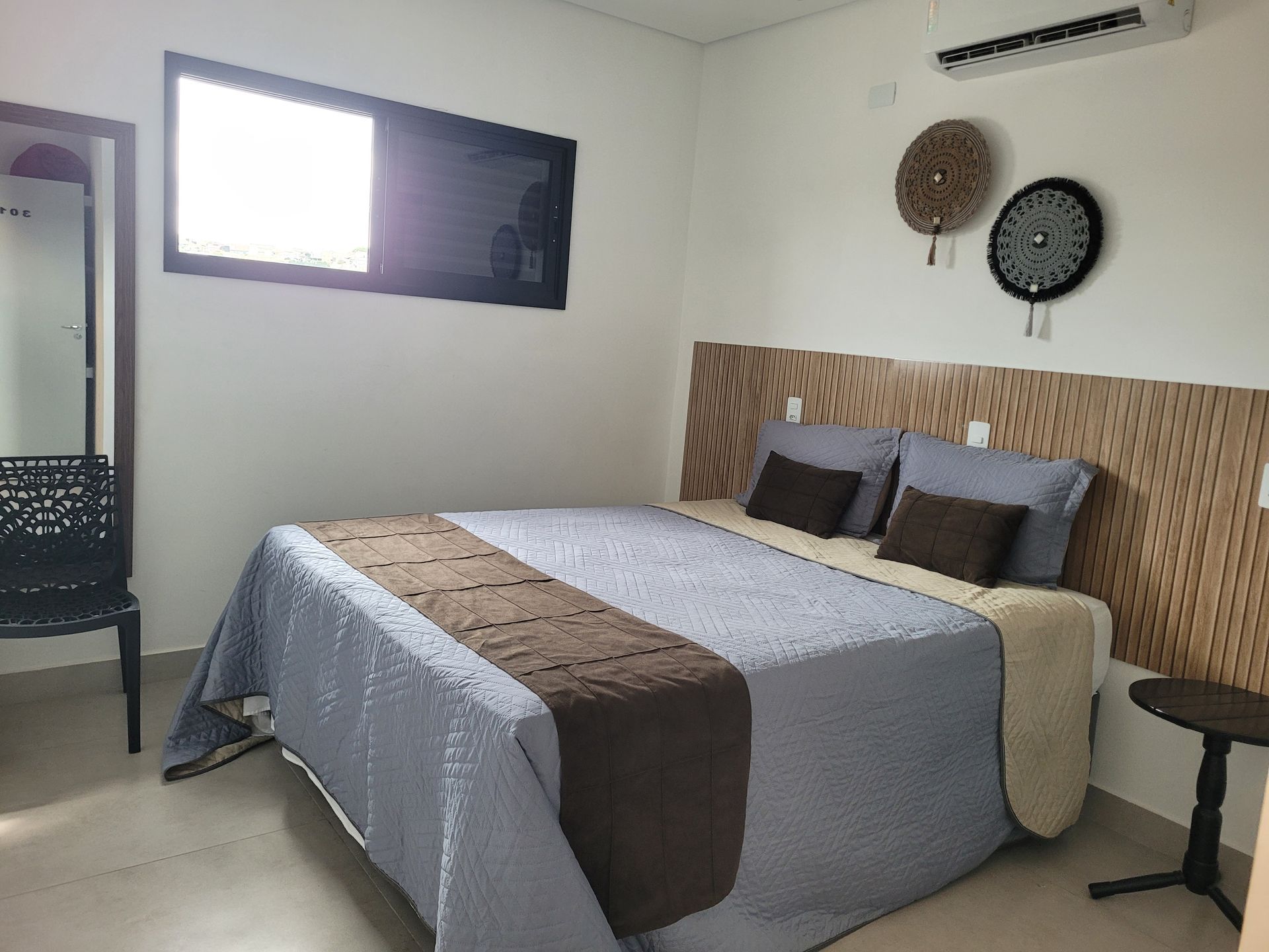 Quarto minimalista com cama, roupa de cama cinza e marrom, espelho de parede e janela.