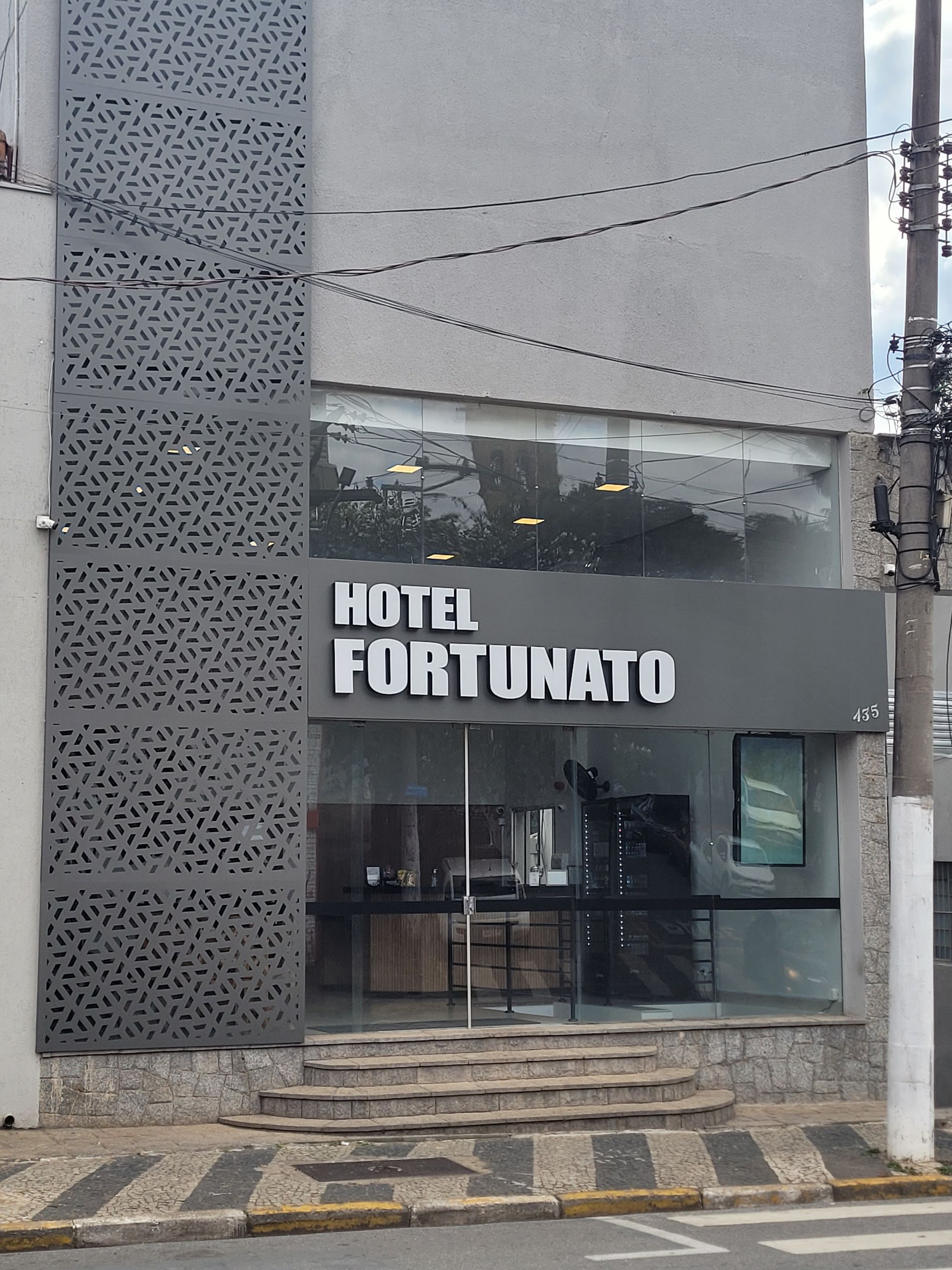 Fachada do Hotel Fortunato com fachada escura, entrada de vidro e painéis de parede estampados em uma rua da cidade.