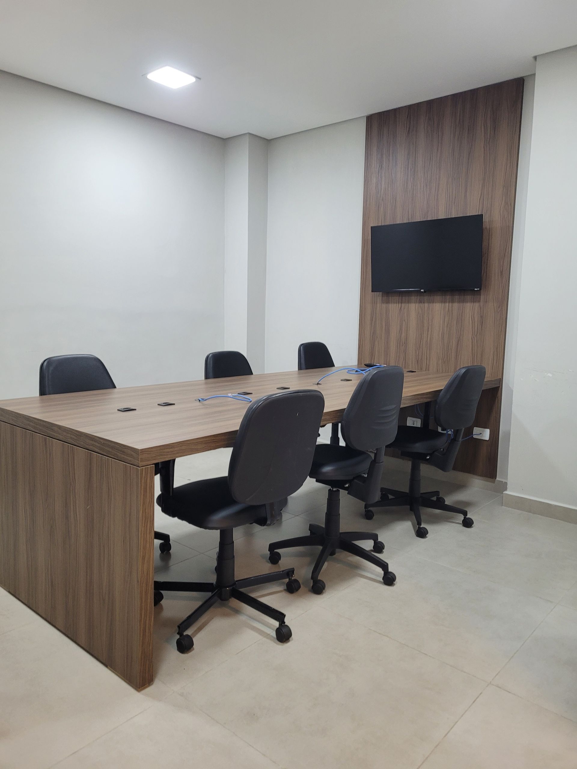 Sala de conferências com mesa de madeira, cadeiras giratórias pretas, TV fixada na parede e painel decorativo de madeira.