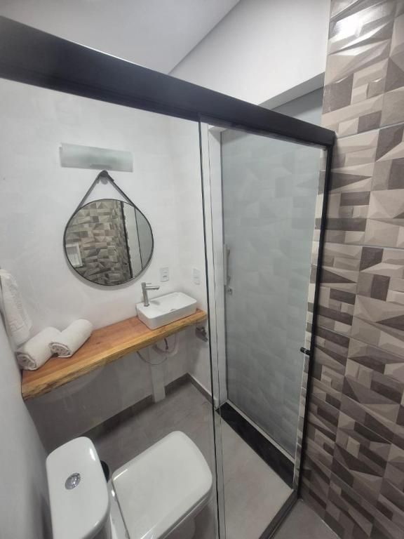 Banheiro pequeno com vaso sanitário, pia, chuveiro e bancada de madeira ao lado de uma porta de vidro fosco.