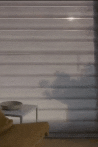 Window with horizontal blinds, sunlight streaming through, casting shadows on a nearby wall and table.
