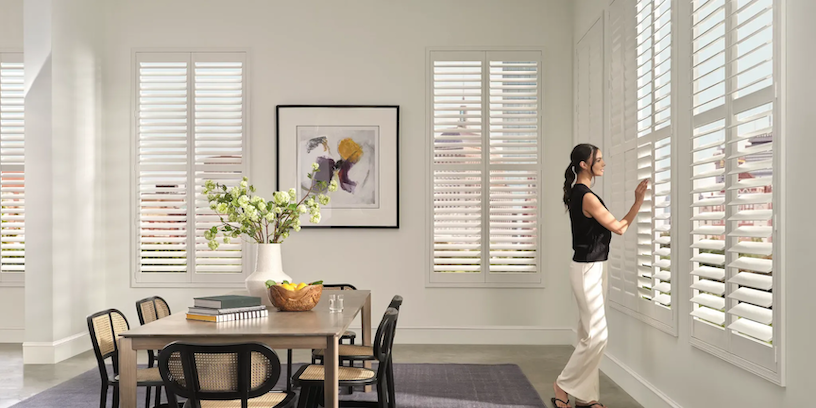 Woman in a dining room looking out of the window with shutters. Dining table, chairs, and art are visible.