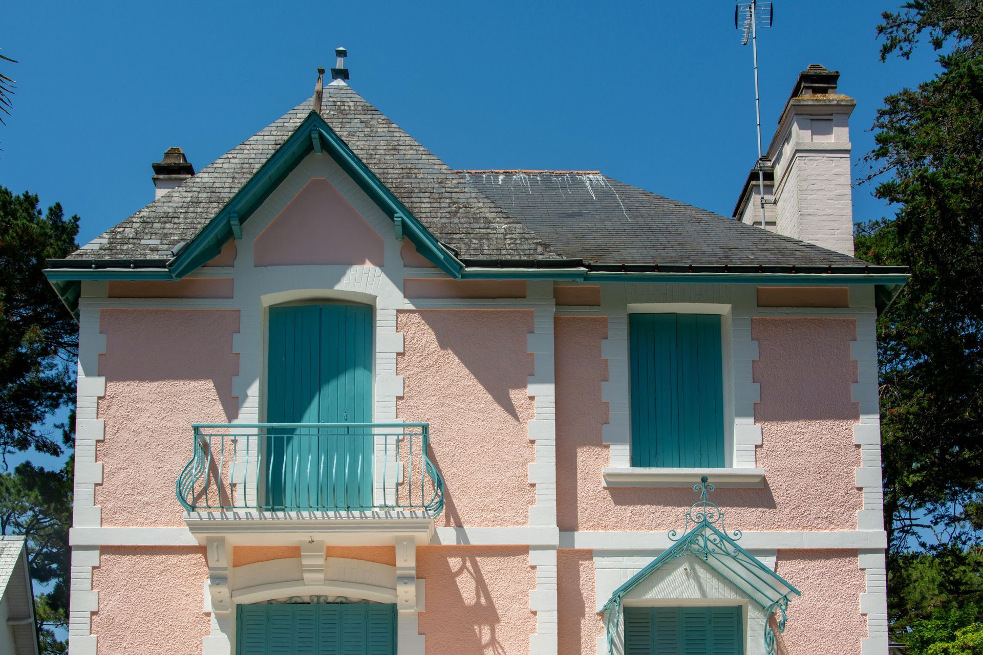 Maison rose avec volets turquoise, balcon et garniture sur fond de ciel bleu.