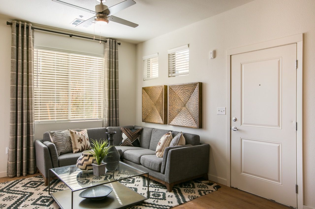 Living room in an apartment with a gray sofa, glass coffee table, and large window blinds.