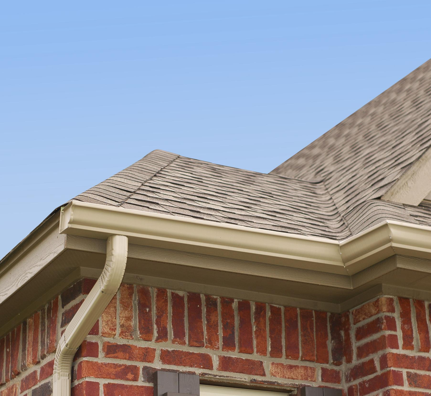 Brick house corner with tan gutters, beige shingle roof, and blue sky.