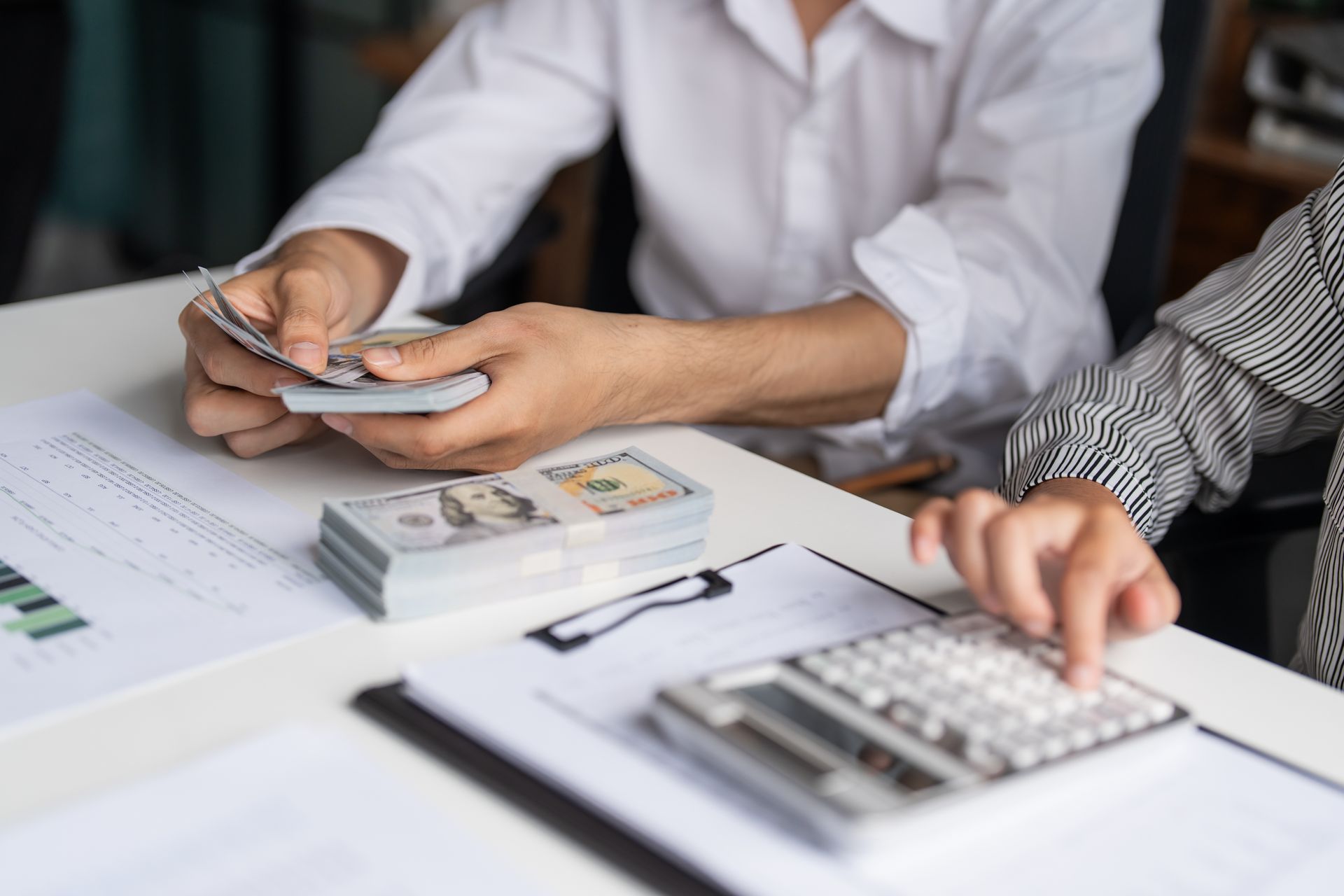 People counting cash and entering figures into a calculator at a desk with documents. People counting cash and entering figures into a calculator at a desk with documents.