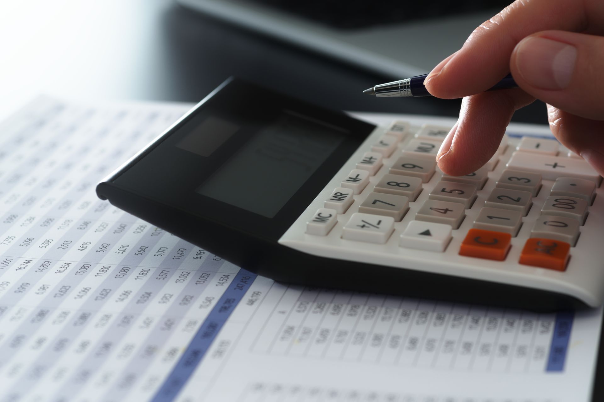 A hand using a white calculator with orange and gray buttons over a printed financial spreadsheet.