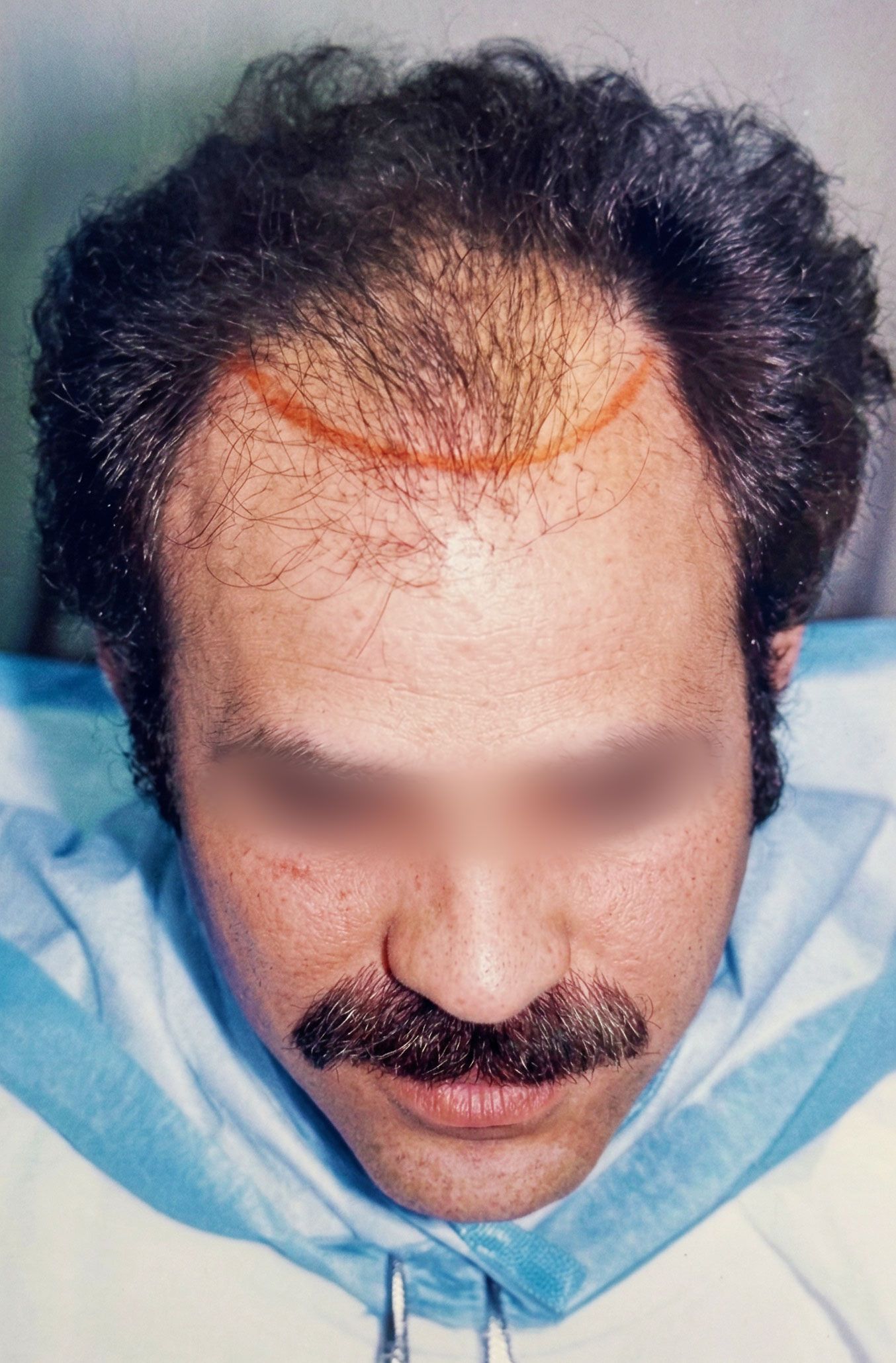 Close-up of a man with a mustache, looking down, wearing a light blue shirt against a plain background