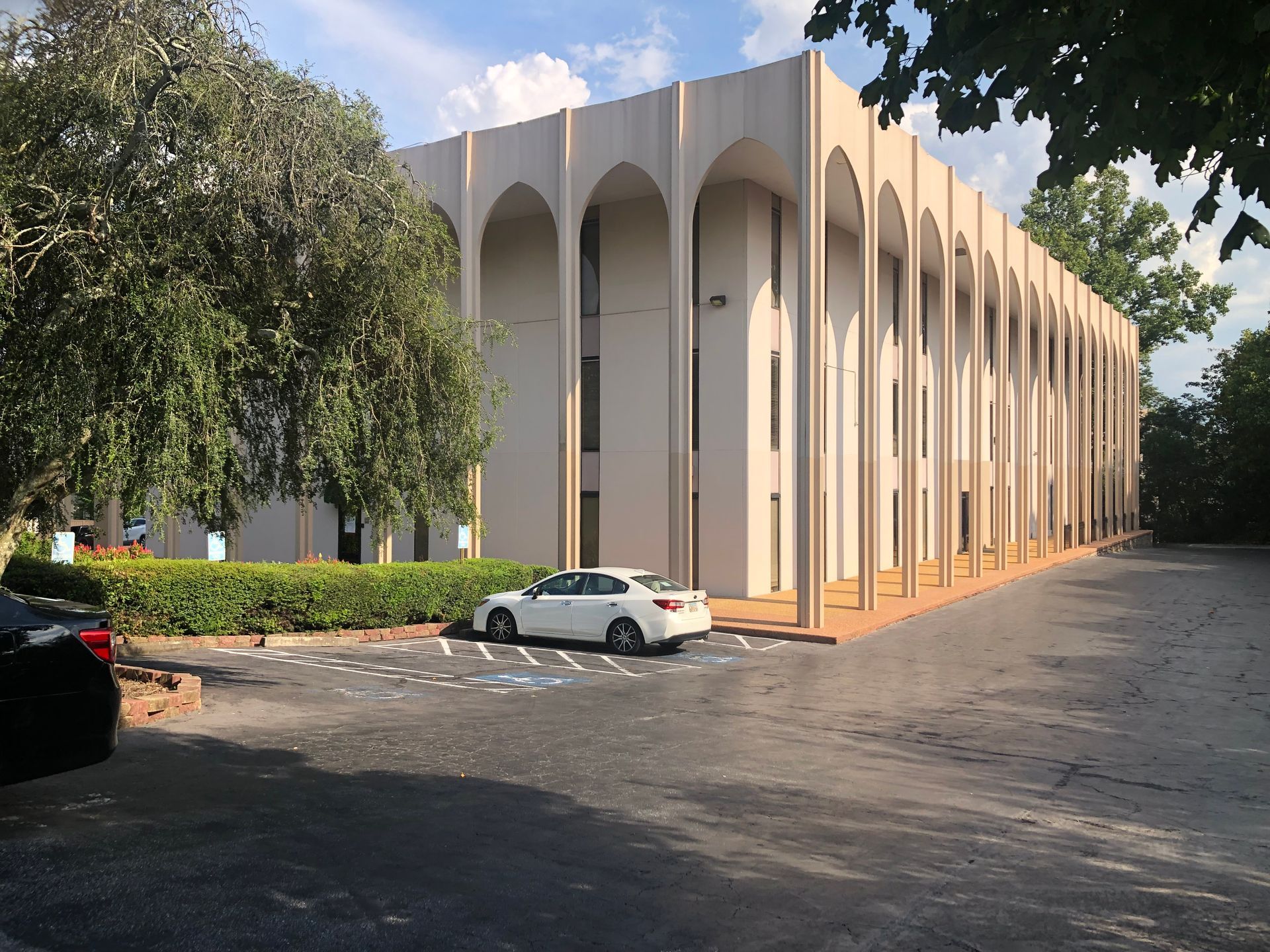 Modern beige building with arched columns beside a parking lot and trees on a sunny day