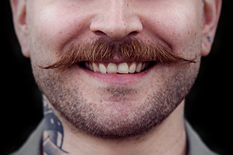 Close-up of a person with a mustache and light facial stubble smiling, with a visible tattoo on their neck.