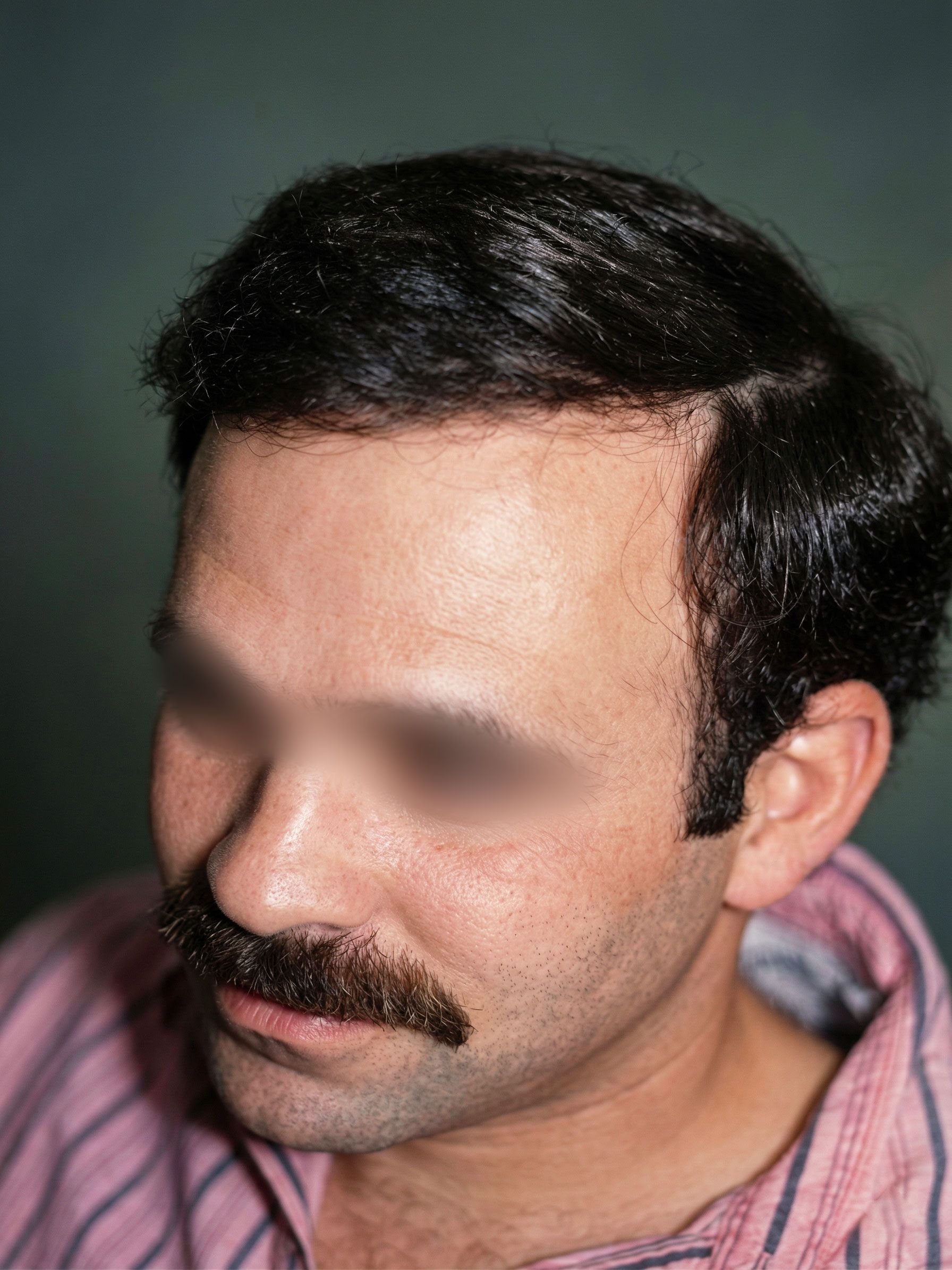 Man with dark hair and mustache in a pink shirt, shown in a close-up portrait against a plain background.