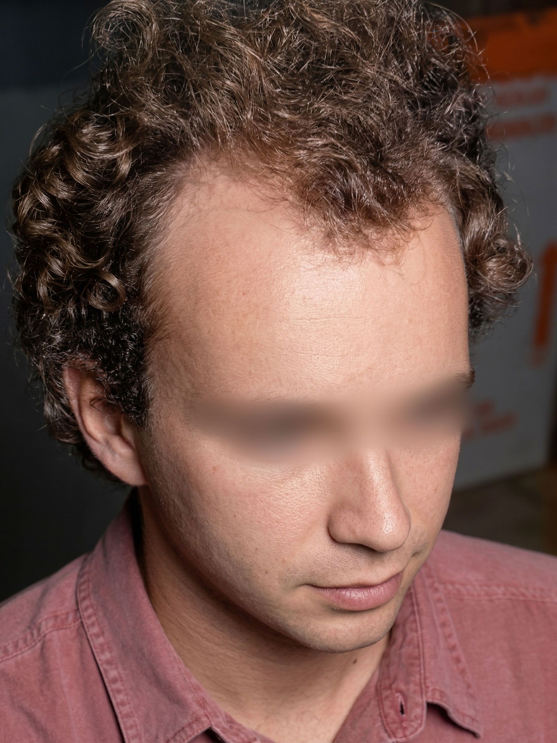 Close-up of a person with curly hair in a pink shirt, looking down in a dim indoor setting