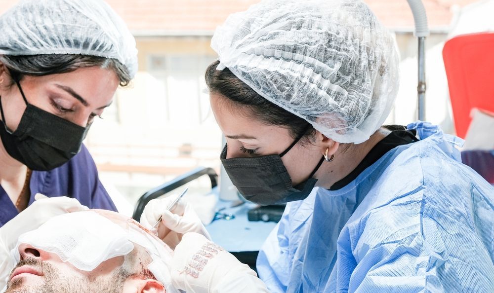 Two medical professionals in scrubs and masks perform a hair transplant procedure on a patient in a clinical setting.