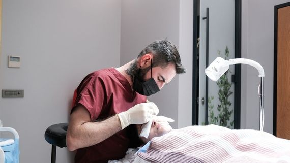 A clinician in a maroon top and mask performs a procedure on a client lying on a treatment table under a desk lamp.