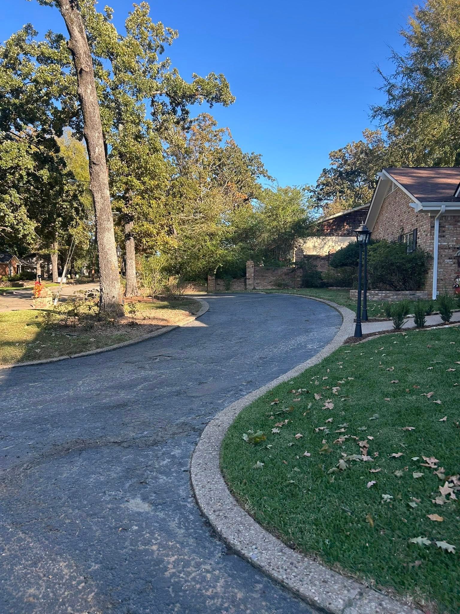 Asphalt driveway curves toward a brick building, bordered by grass and trees under a blue sky.