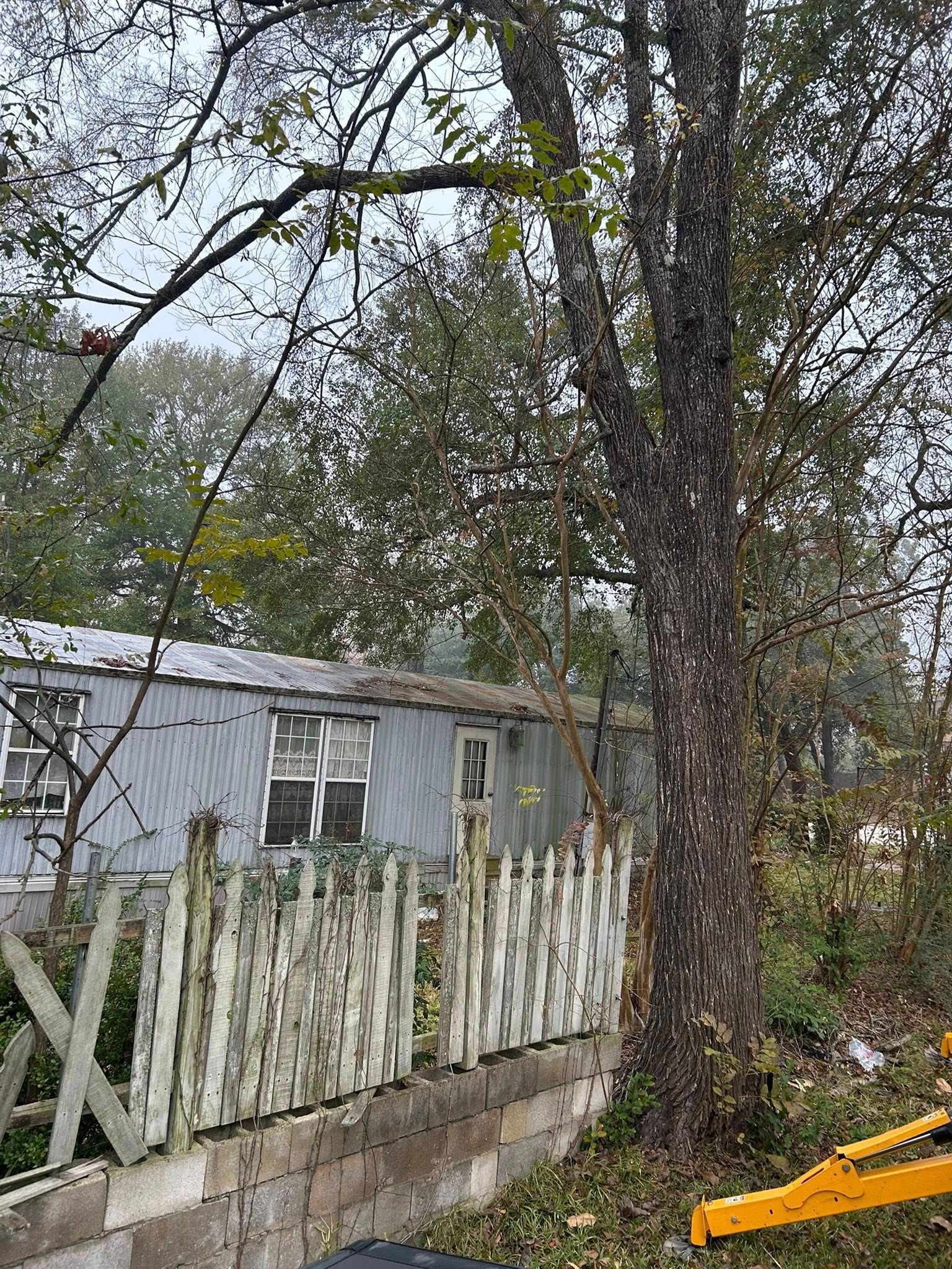 Dilapidated mobile home behind a weathered white picket fence, a large tree to the right, cloudy day.