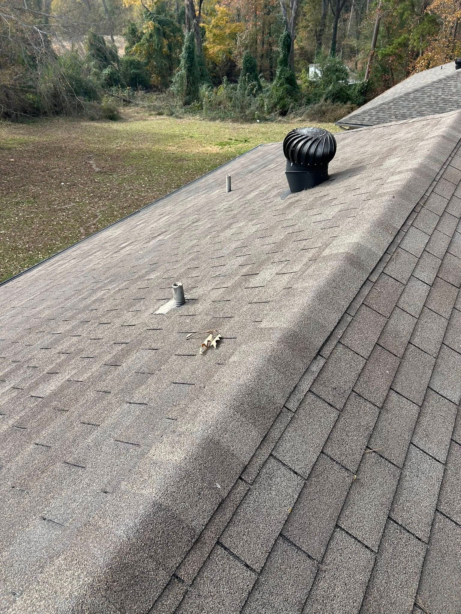 Roof with a spinning vent. Shingles are textured and show some wear. Trees and a yard are in the background.