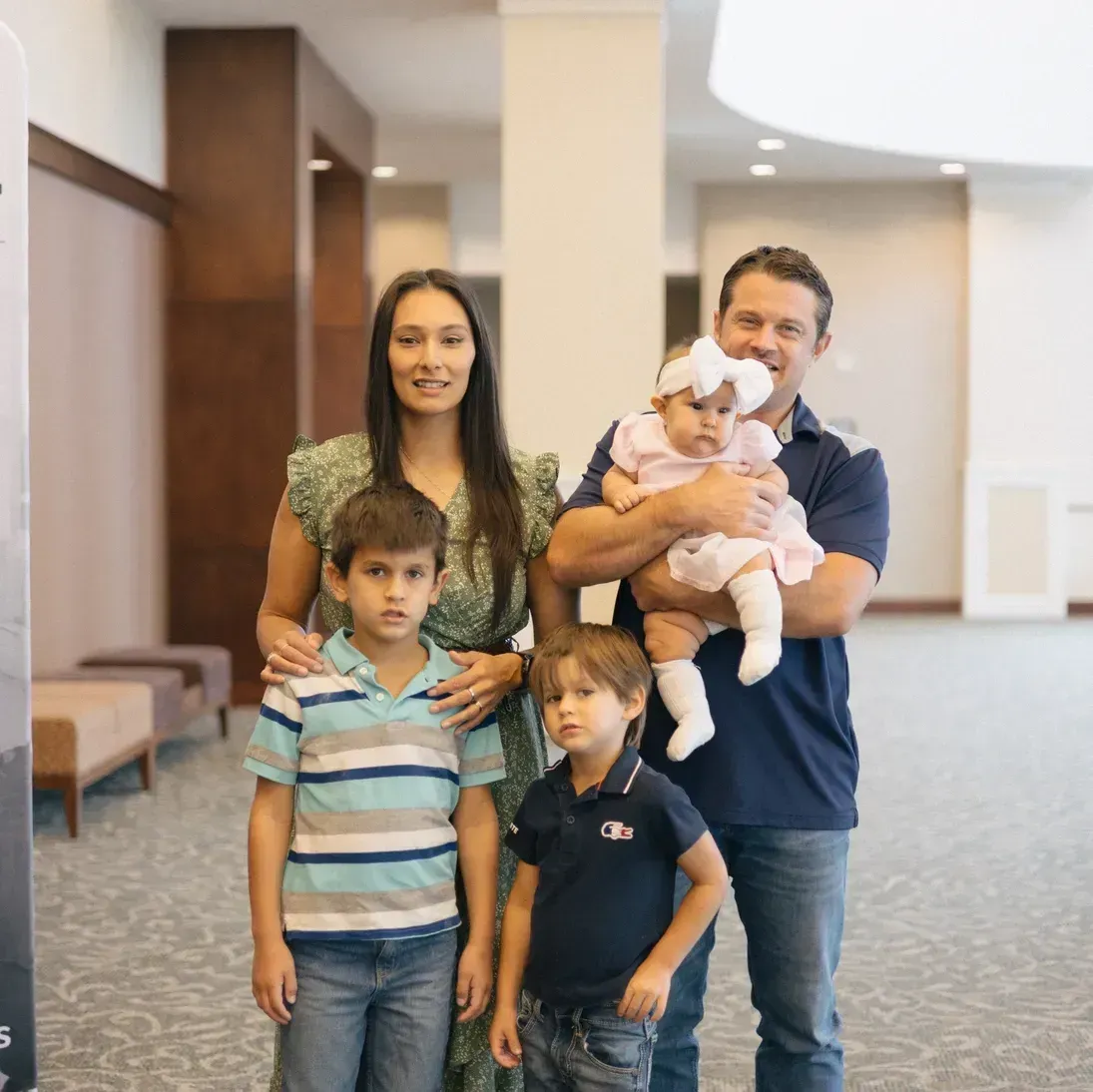 Family of five smiling, indoors: woman, man holding baby, two young boys.
