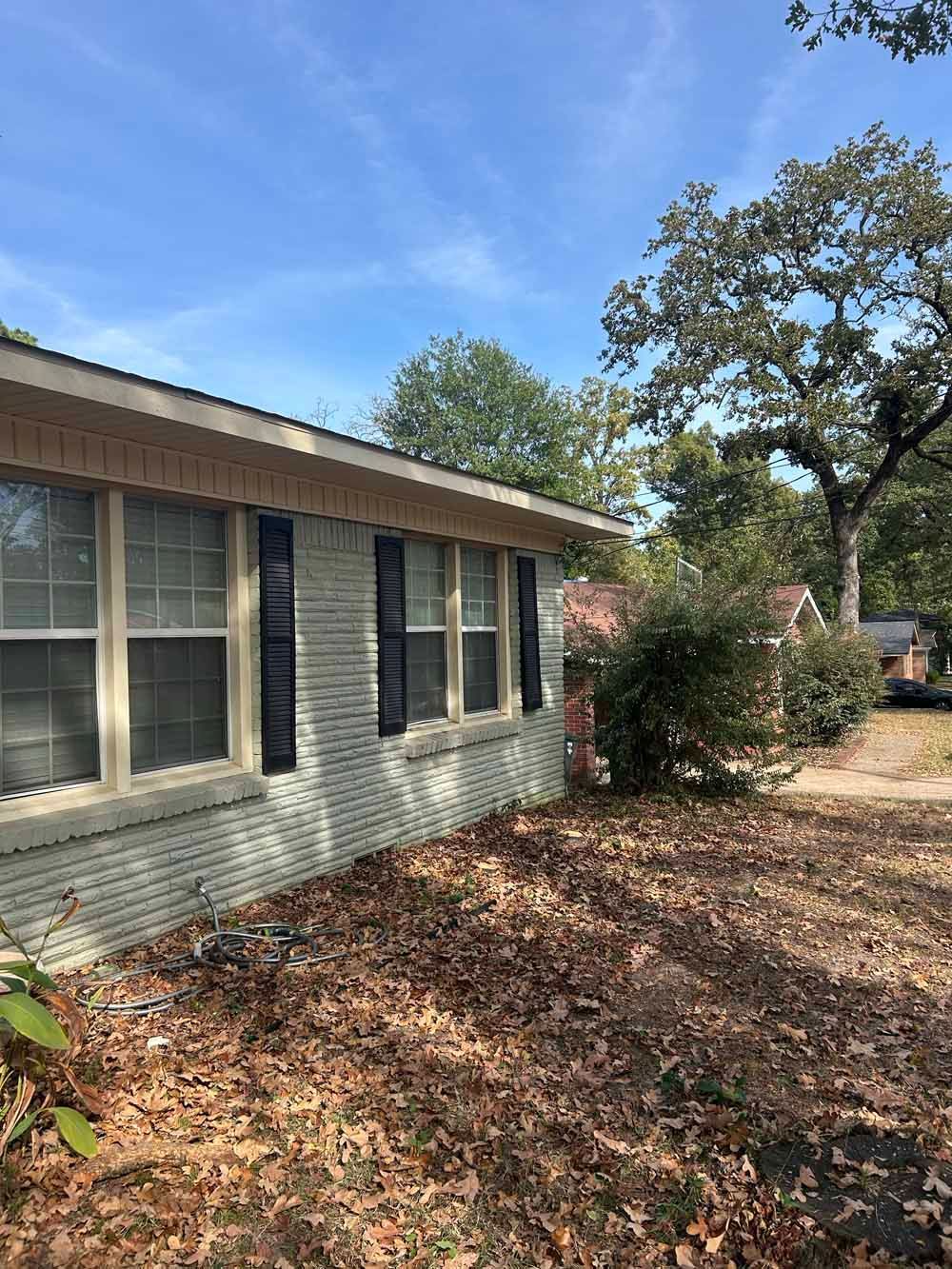 A light green brick house with black shutters, under a blue sky, surrounded by trees and leaves.