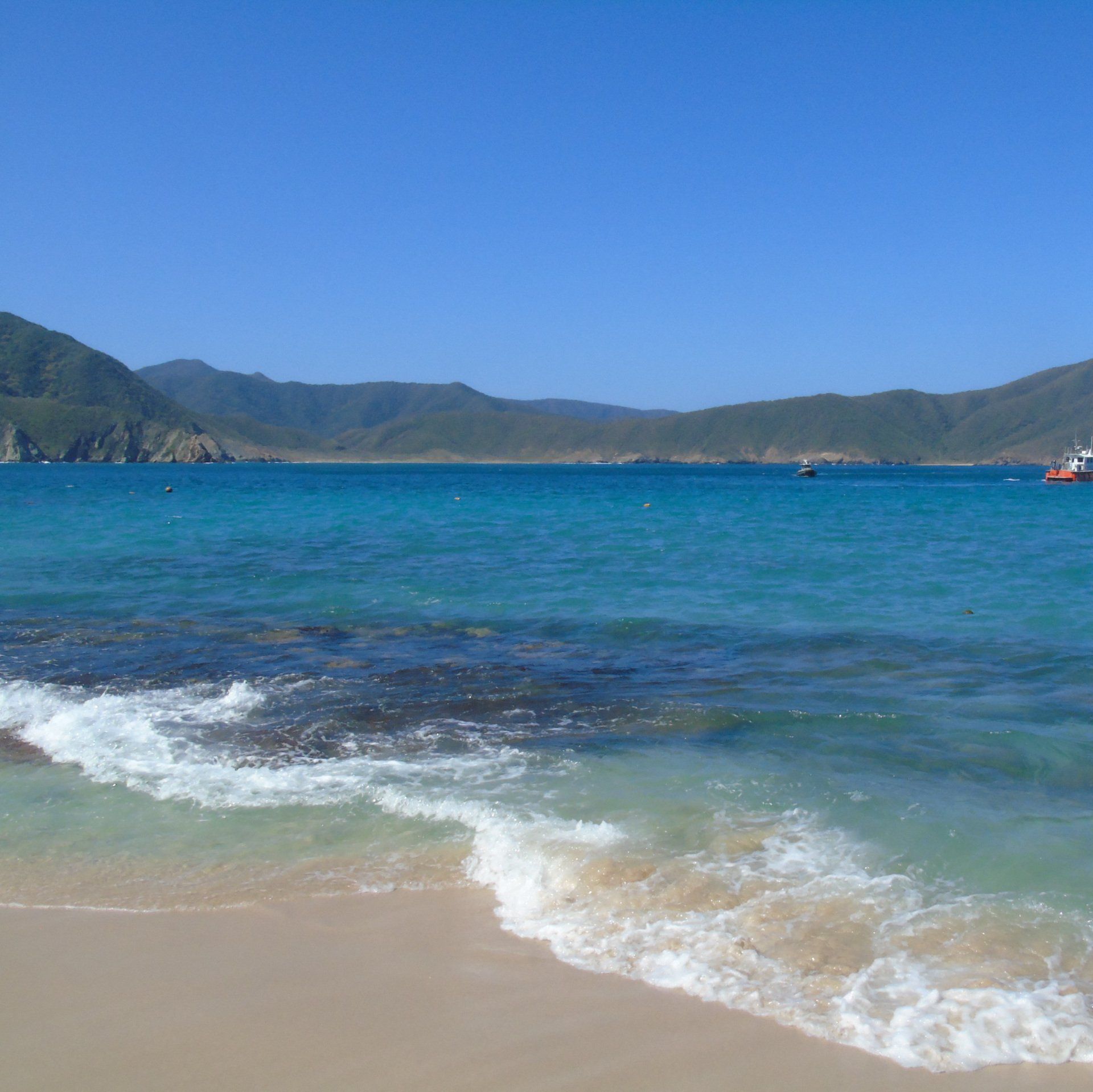 Playa de arena con agua turquesa, montañas al fondo y un cielo azul claro.