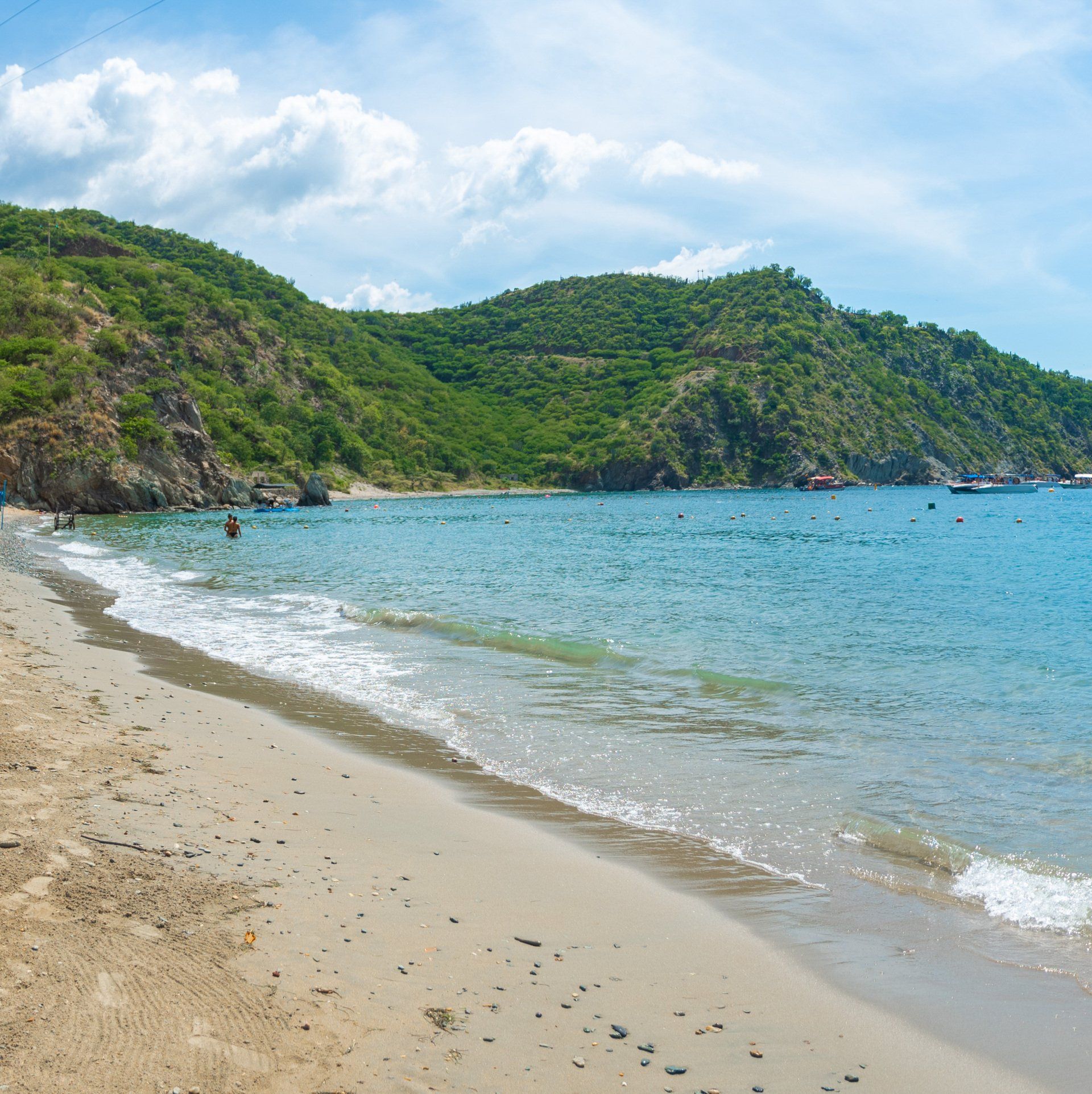 Playa de arena con agua azul clara, montañas cubiertas de vegetación verde y un cielo parcialmente nublado.