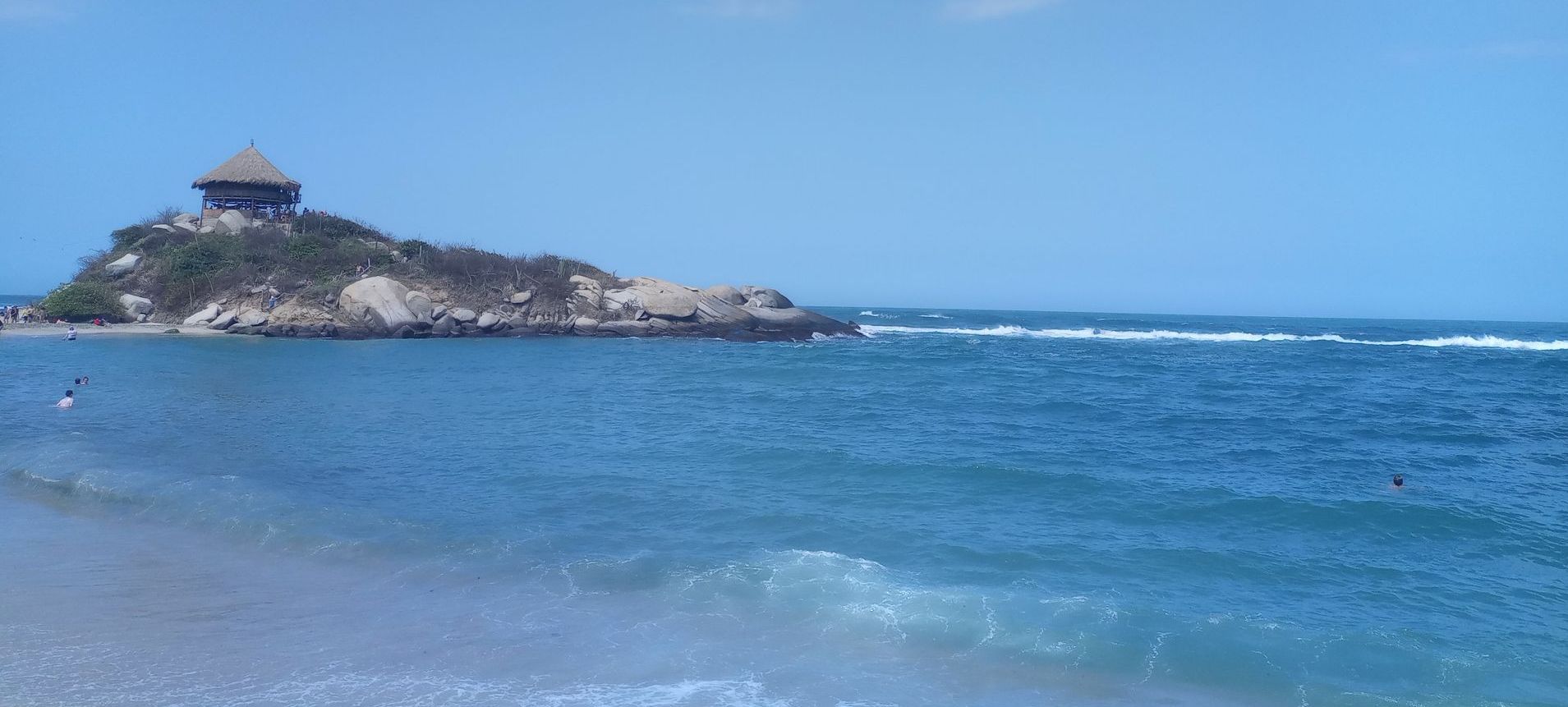 Escena de playa: océano, orilla arenosa, isla rocosa con un pequeño edificio y cielo azul brillante.