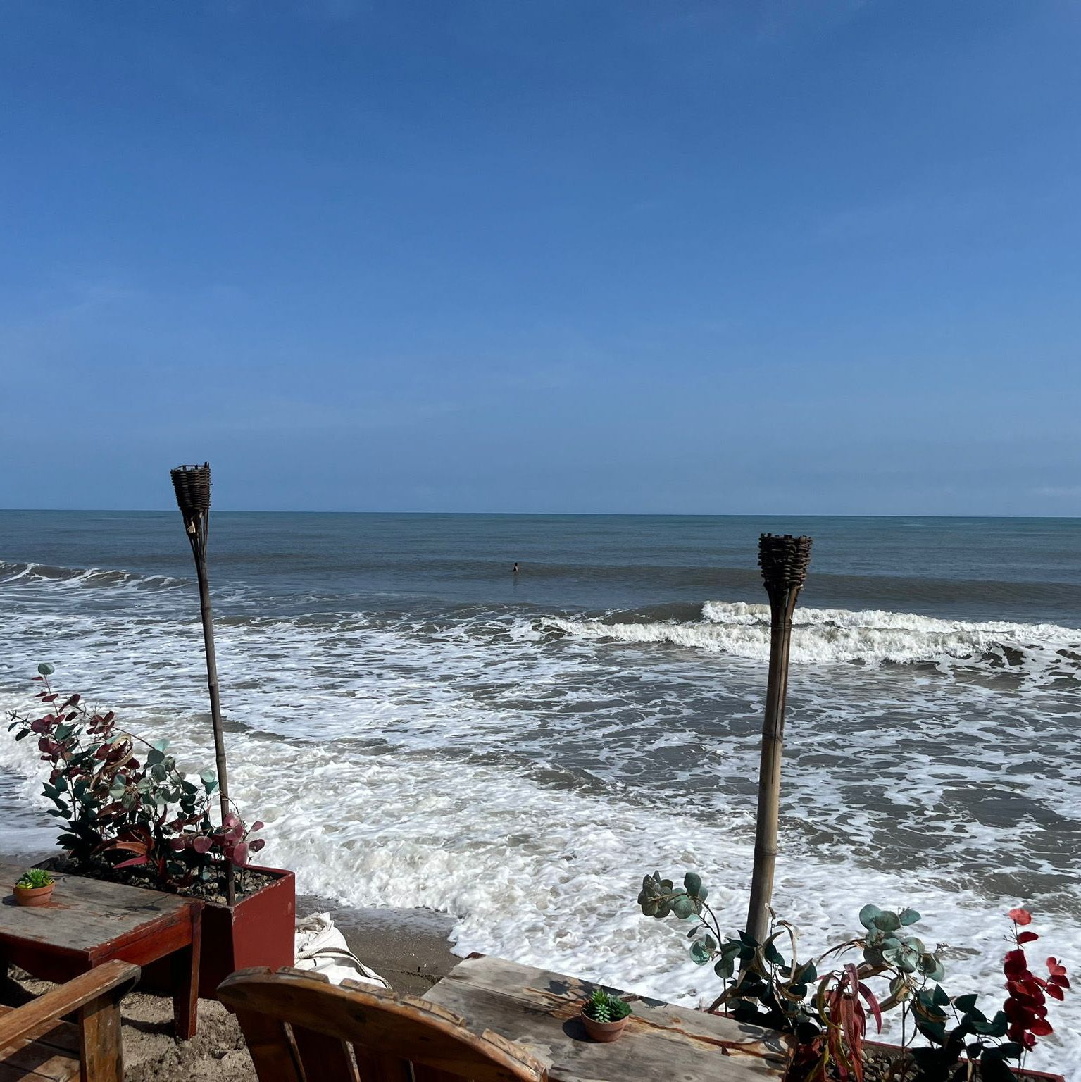 Escena de playa con olas bajo un cielo azul. Antorchas tiki y plantas en macetas flanquean una mesa y una silla de madera.