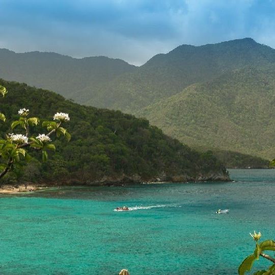 El agua turquesa se encuentra con una exuberante costa verde y montañas. Los barcos salpican el agua bajo un cielo nublado.