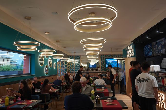 Interno di un ristorante affollato con clienti ai tavoli, pareti color verde acqua e lampade circolari sul soffitto.