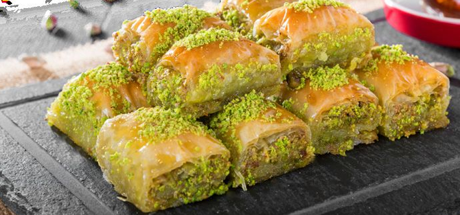 Baklava guarnita con pistacchi tritati, servita su un tagliere di ardesia scura.