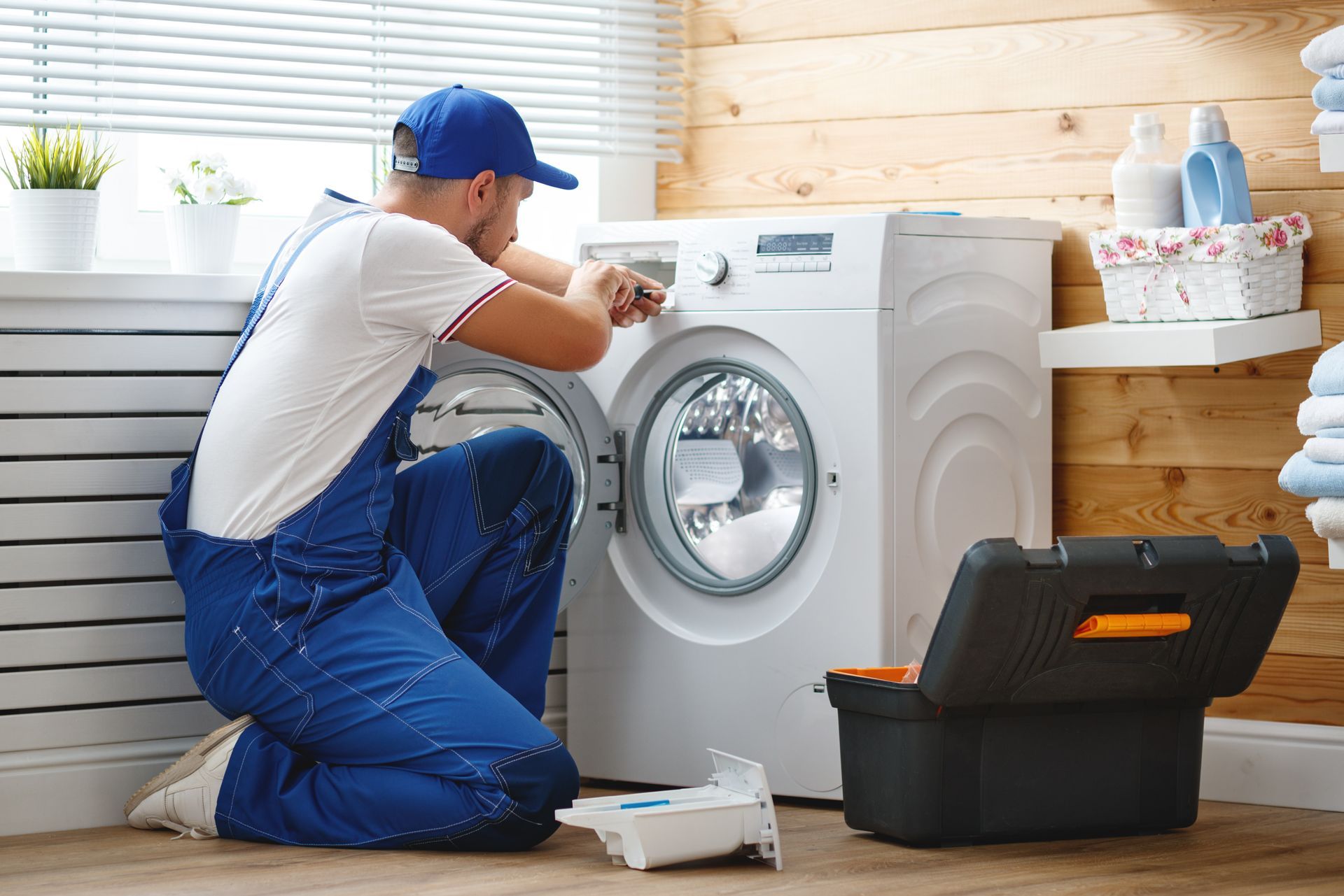 A repair person in blue overalls and cap fixing a white washing machine with a toolbox nearby.