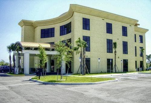 Modern beige office building with curved facade, blue windows, palm trees, and parking lot under clear sky
