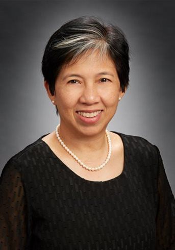 Portrait of a smiling person in a black top and pearl necklace against a gray background