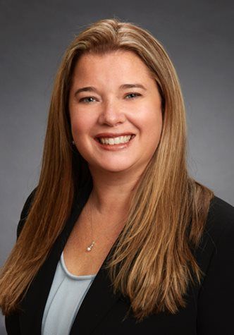 Professional headshot of a smiling woman with long brown hair in a black blazer on a gray background