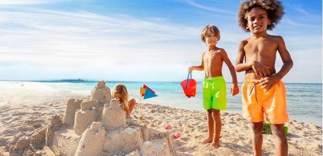 Children on a beach building a sand castle. Blue sky and water behind them.