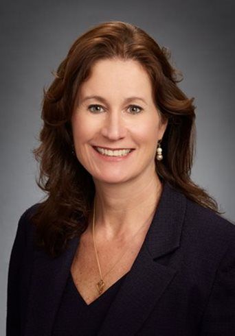 Professional headshot of a smiling woman in a dark blazer against a gray background