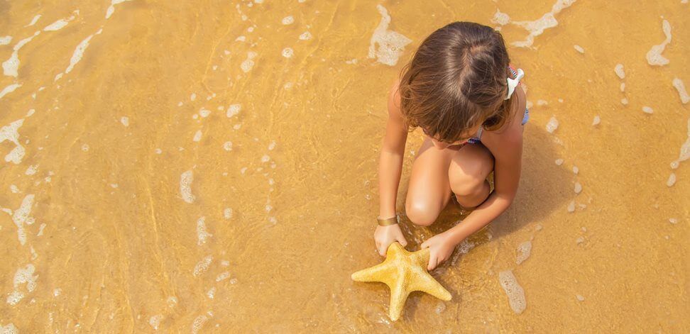 Child on wet sand holding a yellow starfish at the shoreline