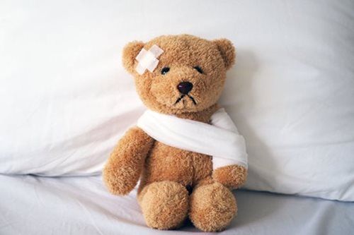 Brown teddy bear with a bandage on its head and a white sling, sitting against a white pillow.