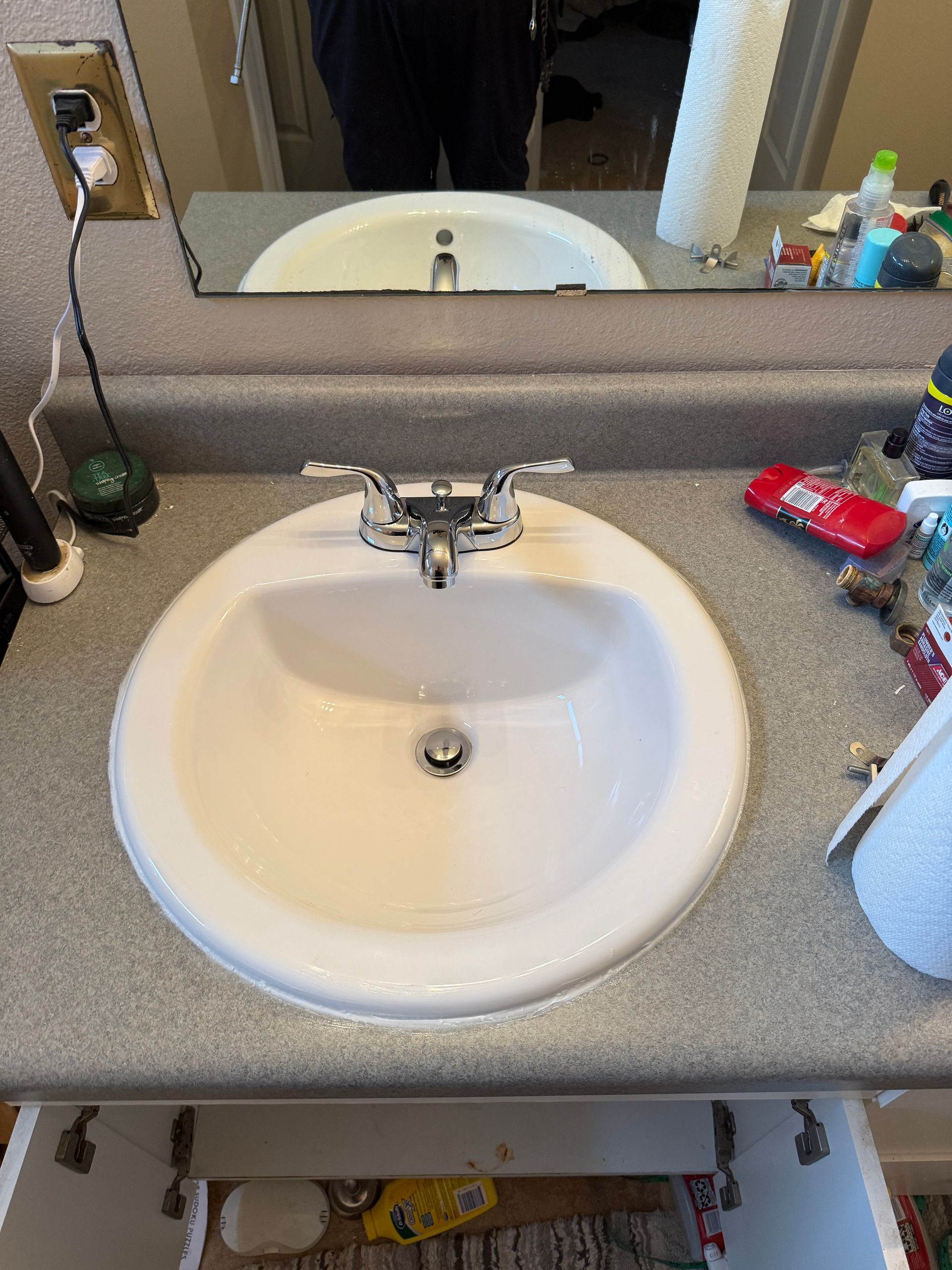 White bathroom sink with chrome faucet. Countertop cluttered with items. Mirror reflects part of a person.