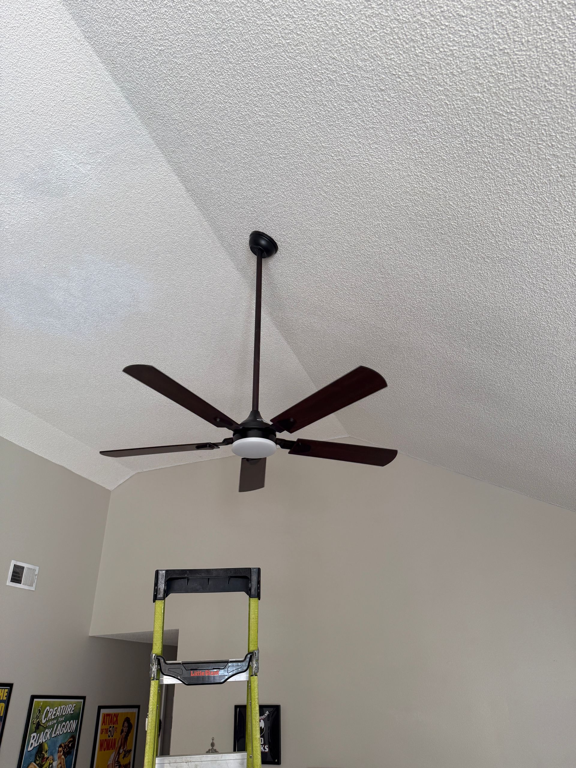 Ceiling fan with five blades hangs from textured ceiling above a stepladder. Beige walls and posters are visible.