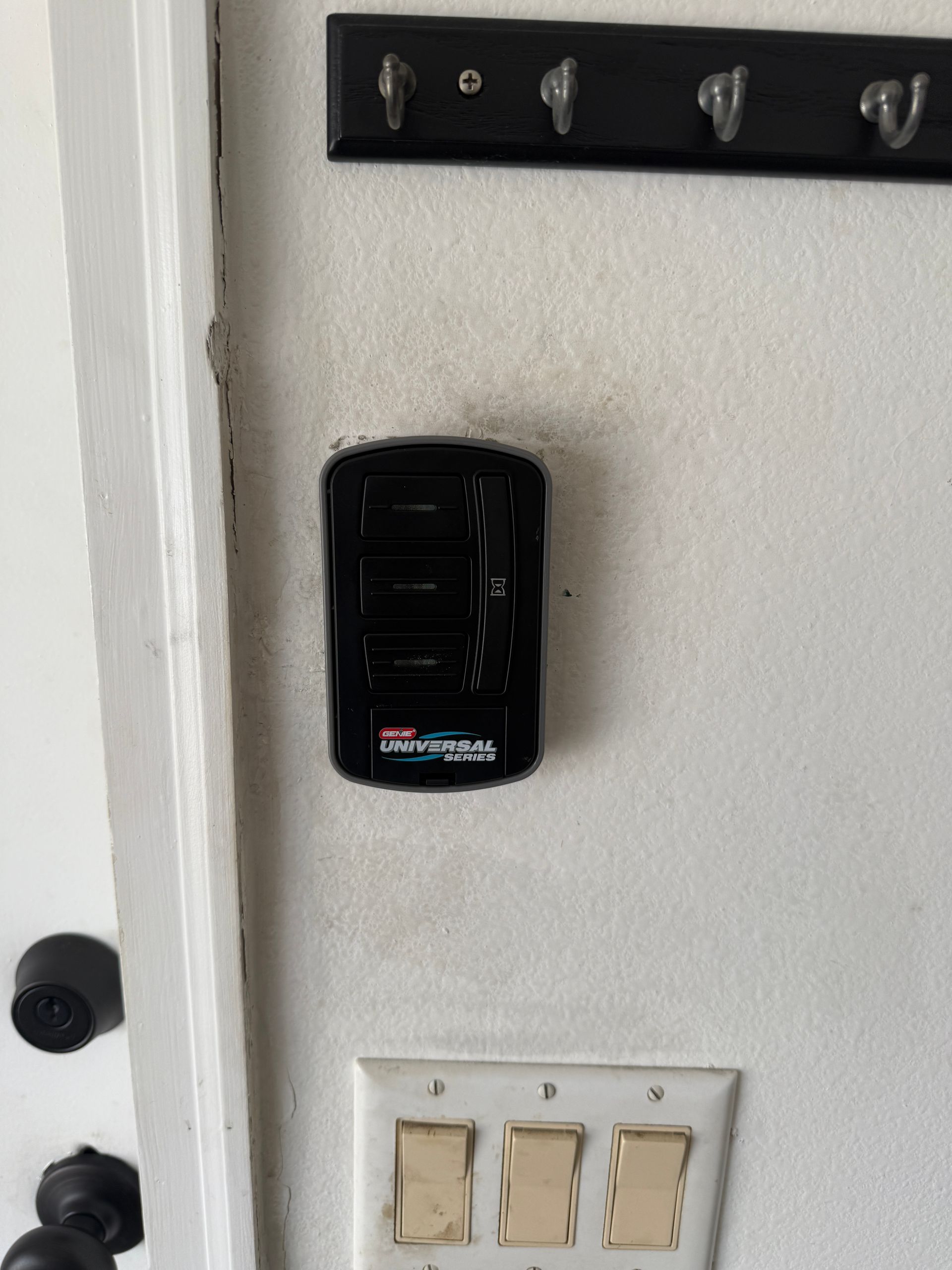 Black garage door opener keypad mounted on a white wall, above light switches and below a key holder.