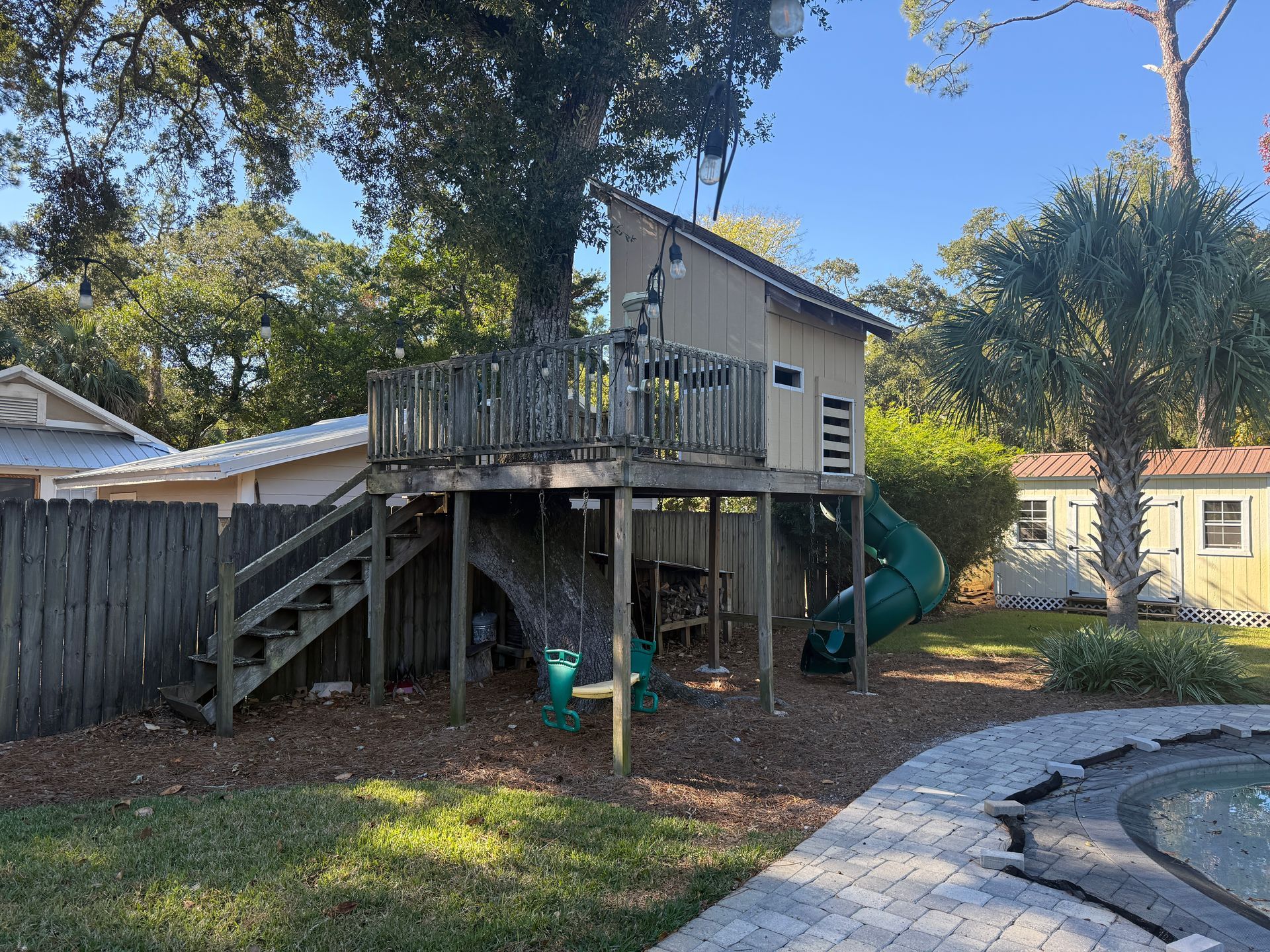 Wooden treehouse with a slide, stairs, and a swing set in a backyard.