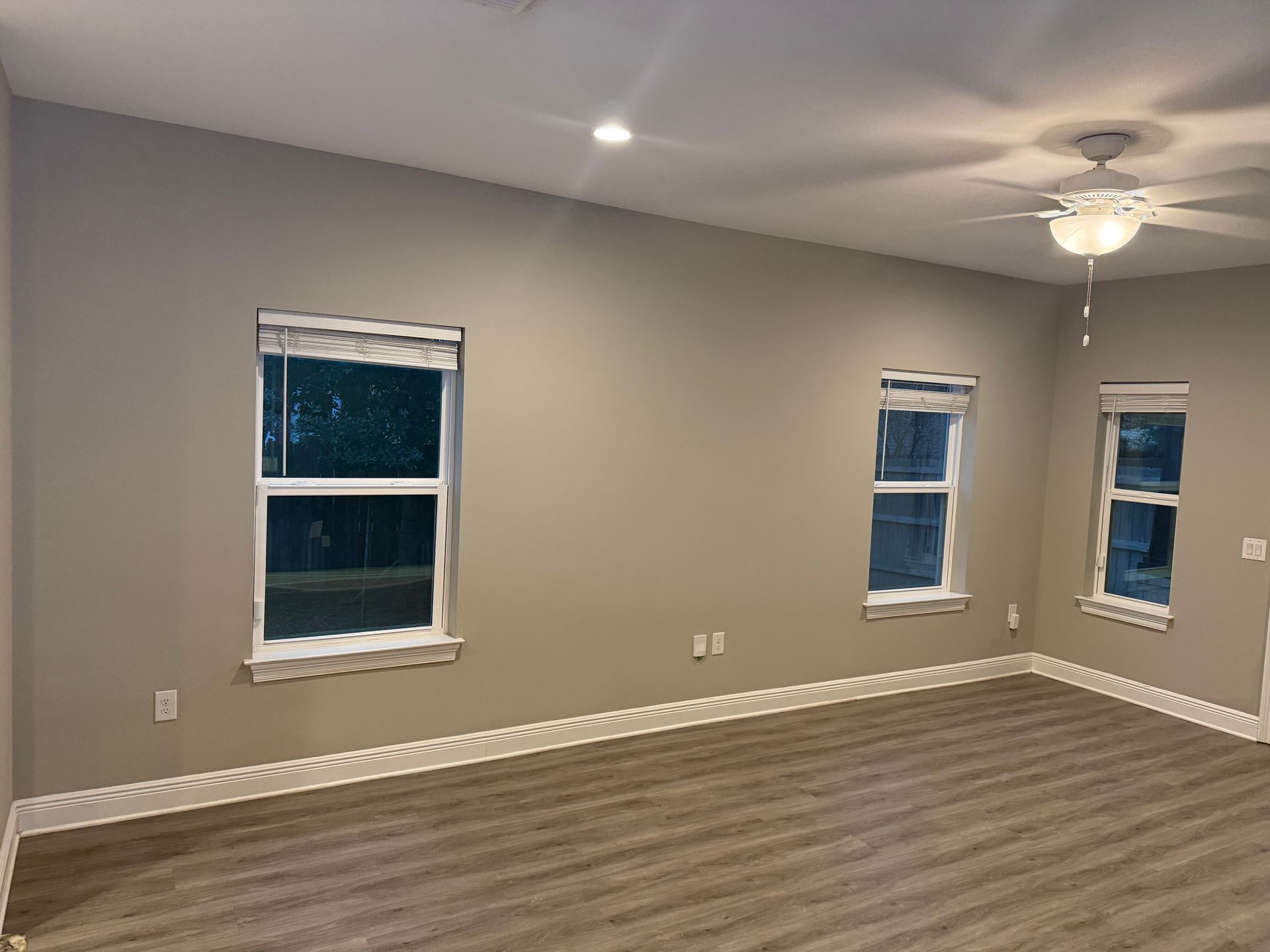 Empty room with three windows, a ceiling fan, and beige walls, with wood-look flooring.