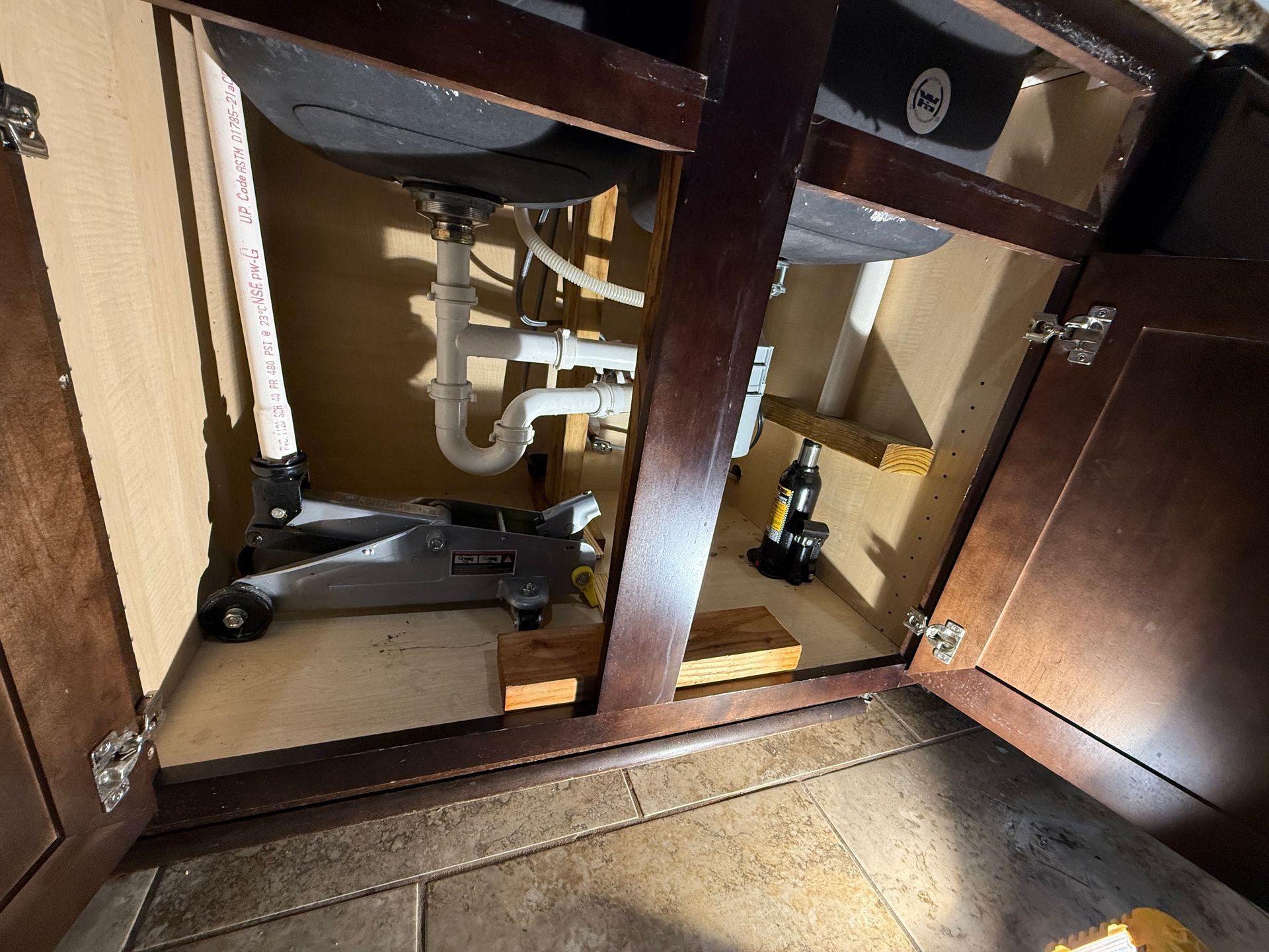 Under-sink cabinet with plumbing visible. A car jack and wooden blocks are used for support.